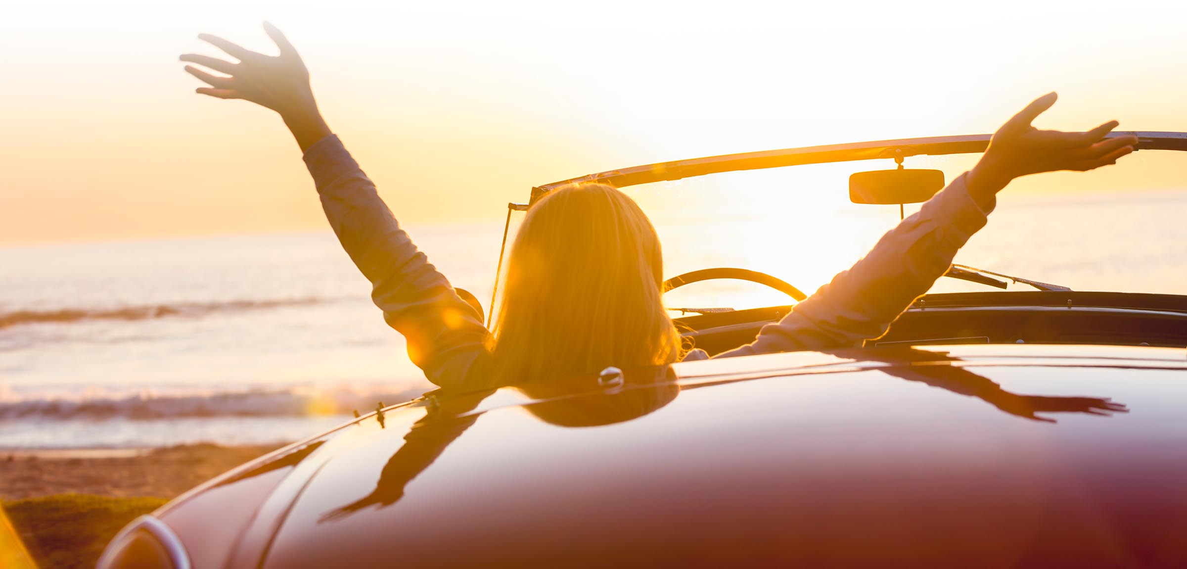 Eine Frau sitzt beim Sonnenuntergang im Cabrio und breitet ihre  Arme aus